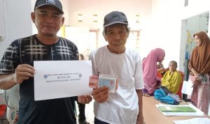 Sangadi Tutuyan Mikdad Modeong mengenakan kaos hitam bergaris putih saat menyerahkan bantuan BLT kepada Keluarga Penerima Manfaat.
