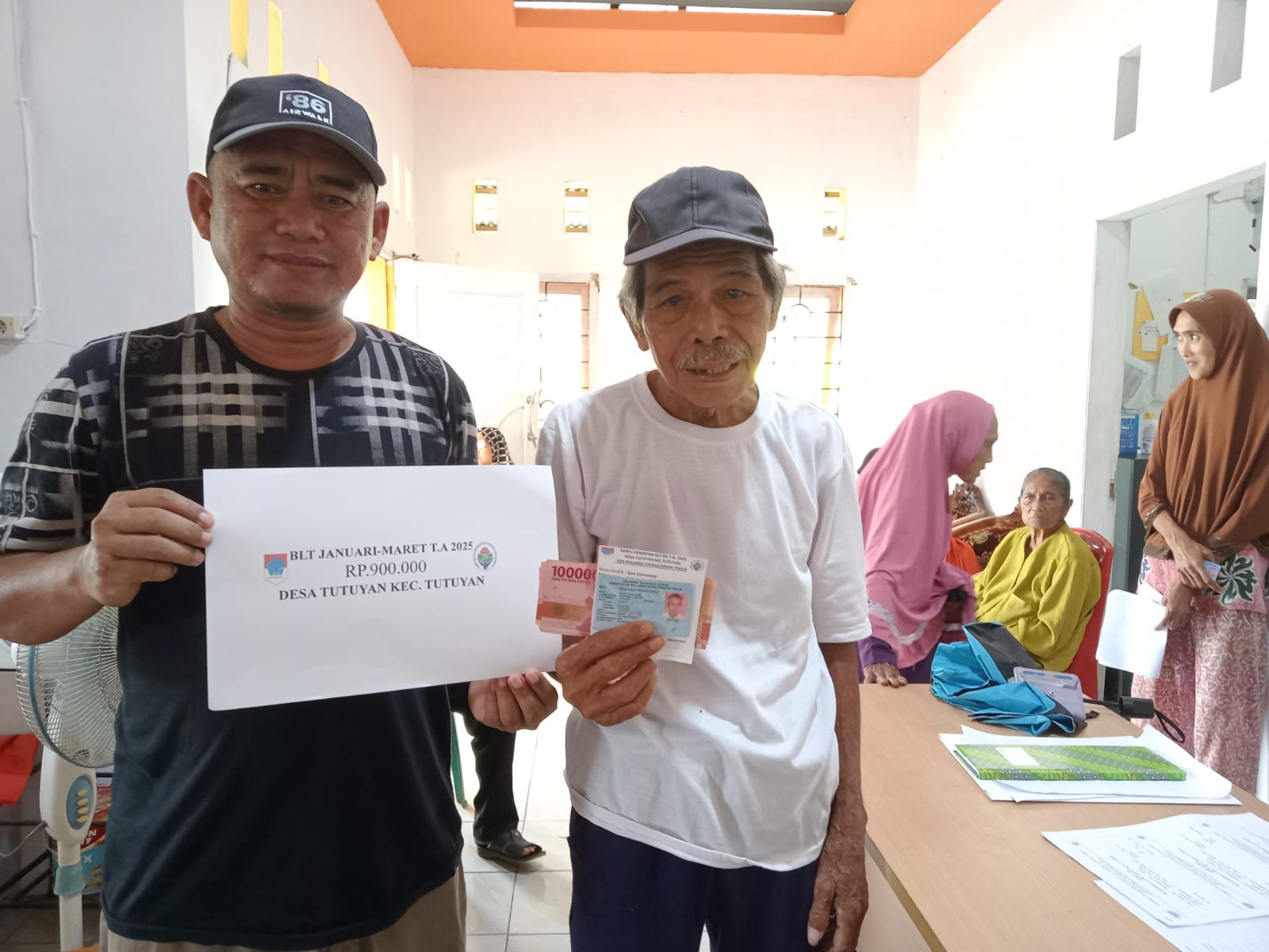 Sangadi Tutuyan Mikdad Modeong mengenakan kaos hitam bergaris putih saat menyerahkan bantuan BLT kepada Keluarga Penerima Manfaat.