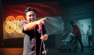 Ramdan Buhang berdiri di depan latar bendera Merah Putih peringatan 80 tahun kemerdekaan. Sebuah ironi, ketika simbol kebanggaan bangsa berkibar megah, pelayanan kesehatan di dalam negeri justru membuat rakyat mencari harapan di negeri orang.