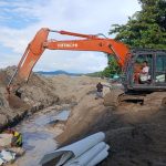 Pekerja proyek bersama alat berat ekskavator menata batu bolder pada pembangunan pengaman pantai di kawasan Batu Pinagut, Desa Boroko Timur, Kecamatan Kaidipang, Kabupaten Bolaang Mongondow Utara. Proyek ini telah mencapai progres fisik 50,05 persen dan berjalan tanpa kendala. (Foto: dok. binadow.com)