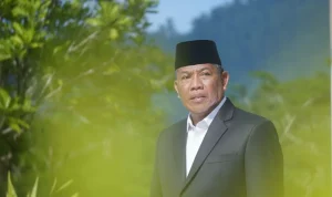 Wakil Ketua DPRD Boltara, Drs. Hi. Depri Pontoh, mengenakan setelan jas hitam dan peci hitam berpose di luar ruangan dengan latar pepohonan hijau.