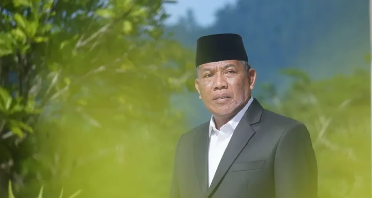 Wakil Ketua DPRD Boltara, Drs. Hi. Depri Pontoh, mengenakan setelan jas hitam dan peci hitam berpose di luar ruangan dengan latar pepohonan hijau.