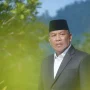 Wakil Ketua DPRD Boltara, Drs. Hi. Depri Pontoh, mengenakan setelan jas hitam dan peci hitam berpose di luar ruangan dengan latar pepohonan hijau.