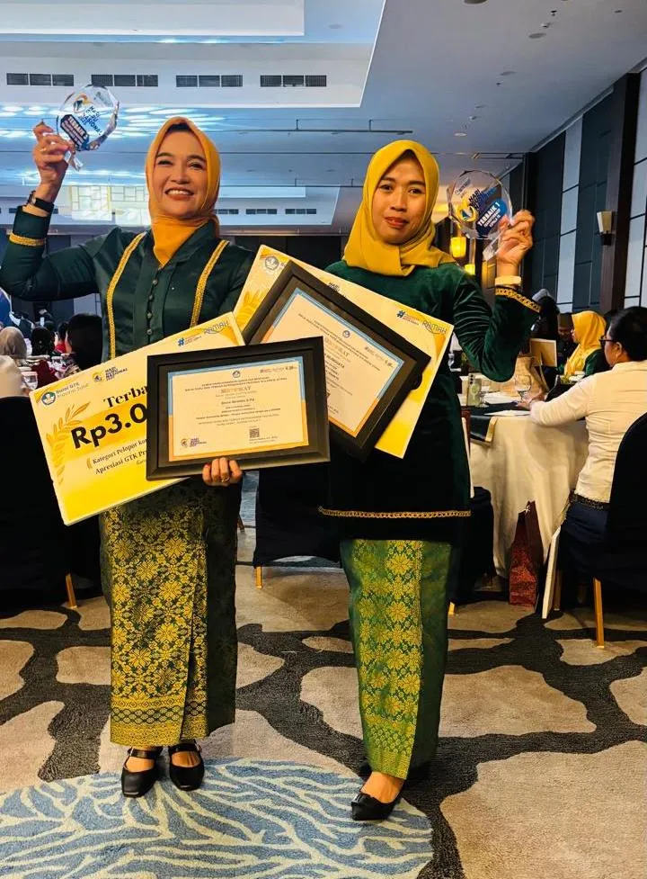 Dua orang guru mengenakan busana adat hijau berpose sambil memegang piala, sertifikat, dan hadiah dalam sebuah ruangan acara.