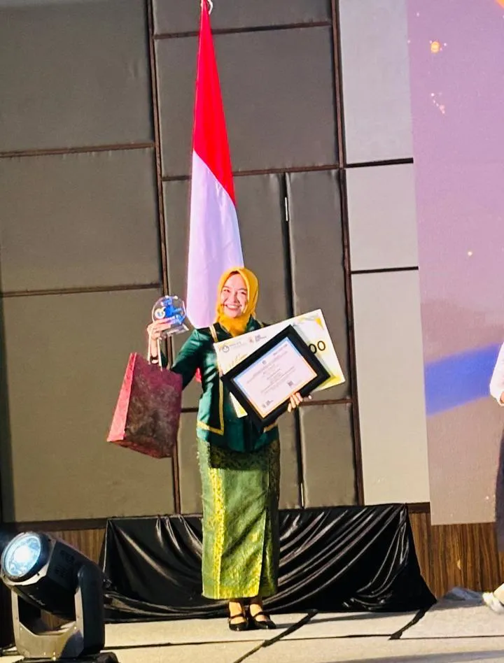 Seorang guru mengenakan busana adat hijau berdiri di atas panggung sambil memegang piala, sertifikat, dan hadiah, dengan latar bendera Indonesia.