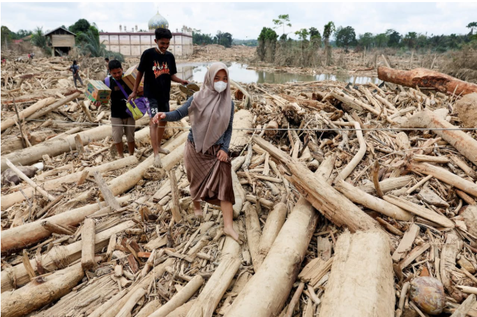 Warga berjalan melewati hamparan kayu gelondongan dan puing banjir bandang di kawasan terdampak Aceh.