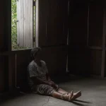Perempuan duduk di lantai rumah kayu dengan kaki terikat kain, sementara seorang petugas mendekatinya untuk melihat kondisi.