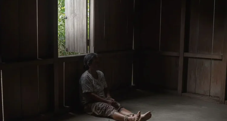Perempuan duduk di lantai rumah kayu dengan kaki terikat kain, sementara seorang petugas mendekatinya untuk melihat kondisi.
